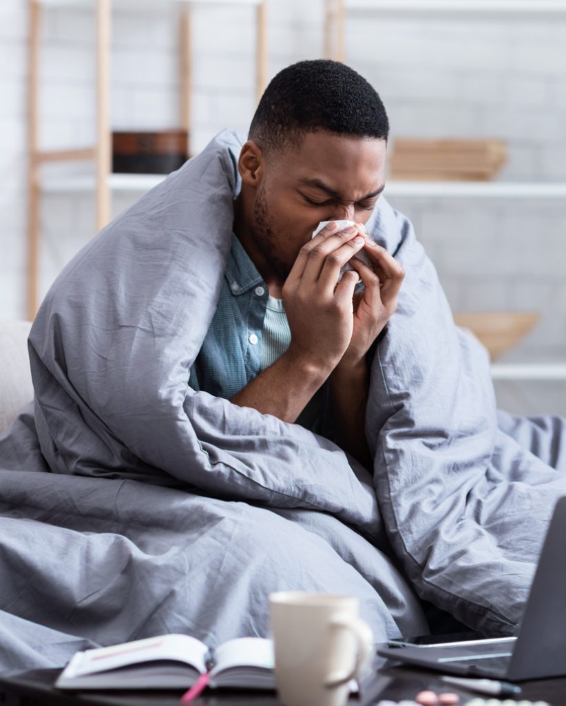 Black man covered with blanket and sneezing into tissue.