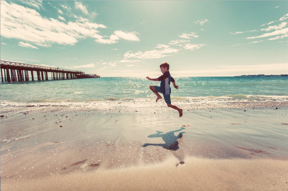 Young boy jumping on the ocean shore by a pier.