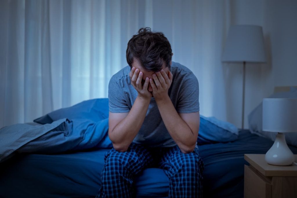 A man sitting on a bed with his face in his hands needs insomnia relief